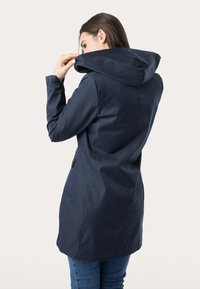 Cappotto blu navy con un tessuto morbido e resistente all'acqua. Presenta una vestibilità leggermente sagomata, tasche laterali e dettagli con bottoni ai polsini.
