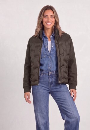 Femme souriante portant une veste matelassée vert olive sur une chemise en denim bleue et un jean bleu taille haute, debout devant un fond uni.