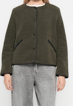 Personne portant une veste polaire texturée vert olive avec des finitions noires et un jean gris délavé, visible des épaules jusqu'aux hanches.