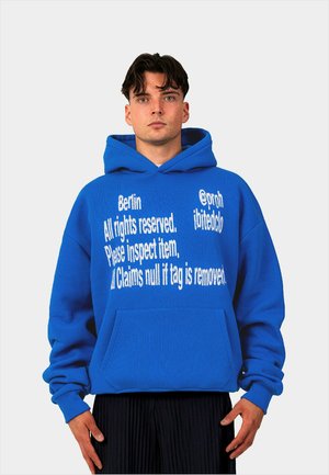 Blauer Hoodie aus dickem Stoff. Mit großem, weißen Text auf der Vorderseite, einschließlich Marke und Prüfhinweisen. Mit Kängurutasche.