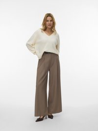 Krémový svetr s výstřihem do V, volný střih; hnědé kalhoty s vysokým pasem a širokými nohavicemi; tmavé lodičky s dekorativními detaily; jednobarevné bílé pozadí.