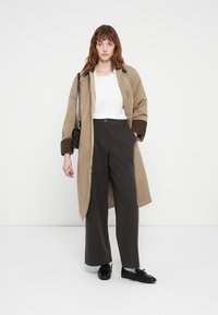 Beige trench coat barna mandzsettákkal, fehér póló, sötétszürke, bővülő nadrág és fekete cipő. A modell egy kicsi, sötét táskába tart.