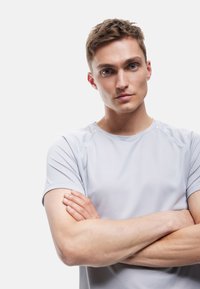 Junger Mann mit kurzen, hellbraunen Haaren und blauen Augen, der ein hellgraues T-Shirt trägt und mit verschränkten Armen vor einem schlichten Hintergrund steht.