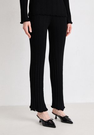 Czarne prążkowane dzianinowe spodnie z falbaniastymi brzegami, noszone z dopasowanym topem z długim rękawem i czarnymi mule na niskim obcasie w kształcie spiczastego czubka, na jasnej podłodze.