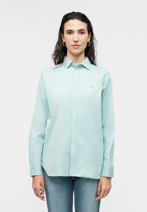 Kvinne med mørkt hår som har på seg gullfargede hoop-øredobber, en lysegrønn stripete skjorte med knapper og blå jeans, stående mot en ensfarget bakgrunn.