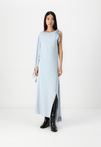 Ανοιχτό μπλε φόρεμα με σχέδιο μίας ώμου και λεπτομέρεια φιόγκου. Διαθέτει μακρύ, ρέον σιλουέτα και πλάγιο σκισίμα, συνδυασμένο με μαύρες μπότες ως το γόνατο.