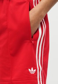 Χέρι τοποθετημένο στην τσέπη με φερμουάρ των κόκκινων σορτς Adidas με λευκές τρεις ρίγες και το λογότυπο Trefoil στο πλάι.