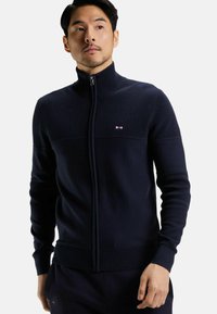 Sweat à zip navy avec texture côtelée et col montant. Présente un logo sur la poitrine et des poignets ajustés pour un look épuré.
