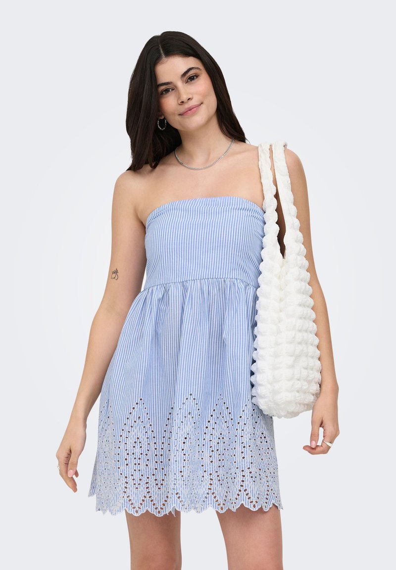 Jeune femme portant une robe rayée bleue et blanche sans bretelles avec un ourlet ajouré, portant un sac à épaule blanc texturé, souriant légèrement.