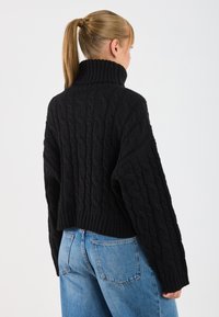 Maglione nero a coste con colletto alto, lunghezza cropped e maniche larghe. Indossato con jeans a vita alta di colore blu chiaro, che mostrano un tessuto testurizzato.