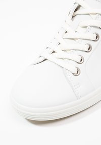 Primo piano di una sneaker bianca con occhielli metallici e lacci bianchi legati strettamente su uno sfondo bianco.