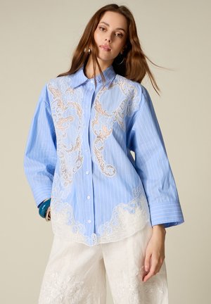 Młoda kobieta ubrana w luźną niebieską koszulę w cienkie paski z misternymi białymi i beżowymi koronkowymi wzorami oraz białe haftowane spodnie o szerokich nogawkach.