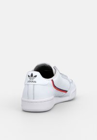 White Adidas sneaker with black trefoil logo on heel, red and blue stripe detail on side, angled to show back and side view.