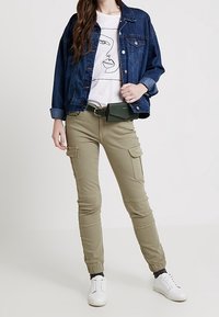 Chaqueta de mezclilla sobre una camiseta gráfica blanca, pantalones cargo en verde oliva y zapatillas blancas. Las características incluyen puños elásticos y bolsillos.