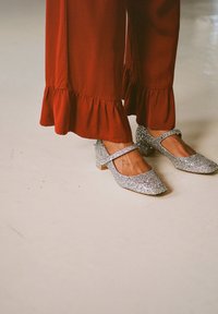 Sidabrinės blizgios Mary Jane bateliai su žemu blokiniu kulnu, turintys kulkšnies dirželį ir apvalią nosį, derinami su raudona suknele su volangais.