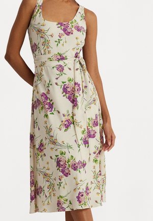 Bez rękawów, kremowa sukienka midi z fioletowym kwiatowym wzorem i przewiązaną w talii szarfą, noszona przez osobę stojącą na białym tle.