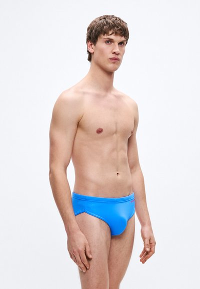 Μπλε μαγιό σλιπ με λεία, γυαλιστερή υφή και ελαστική μέση. Διαθέτει ελάχιστες ραφές και μικρό λογότυπο στο πλάι.