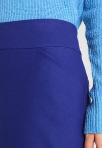 Pull en tricot côtelé bleu associé à une jupe violette unie avec une texture lisse et des poches latérales en biais.