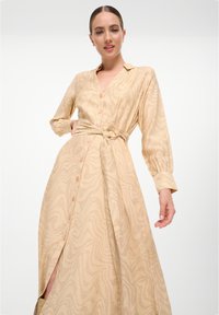 Robe beige à manches longues avec un devant à boutons, une taille ceinturée et un motif texturé ondulé. Présente des poignets et une fente haute sur la cuisse.