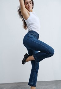 Jeans bleu foncé à coupe slim, avec un ourlet effiloché et une taille haute. Associés à un haut sans manches blanc et des chaussures noires. Texture en denim lisse.
