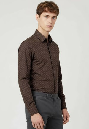 Junger Mann mit braun gemustertem Langarmhemd und grauer Hose, steht vor hellem, einfarbigem Hintergrund und blickt leicht nach links.