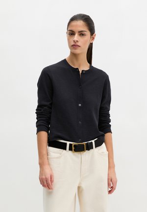 Frau mit dunklem Haar im Pferdeschwanz, trägt eine schwarze, geknöpfte Strickjacke, weiße Hose und einen schwarzen Gürtel mit goldener Schnalle vor einfarbigem Hintergrund.