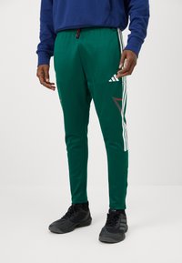 Férfi zöld Adidas tréningnadrágban, fehér csíkokkal és fekete sportcipőben áll egy sima világos háttér előtt.