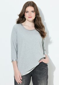 Junge Frau mit langen braunen Haaren, die ein hellgraues Langarmshirt und dunkle graue Jeans trägt, mit der Hand in der Tasche, vor einem weißen Hintergrund stehend.
