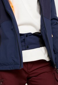 Giacca blu navy con sezioni a contrasto, foderata in nero e con chiusure a scatto visibili, indossata sopra un top in pile bianco e pantaloni bordeaux.