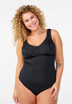 Krøllet kvinde smilende, iført en sort heldragt, stående mod en ensartet hvid baggrund med en rosetatovering på armen.