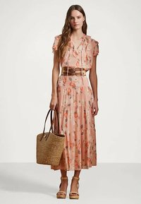 Kvinne iført en rosa blomstret kjole med volanger, et bredt brunt belte, brune platåsandaler og holder en stor vevet totebag.