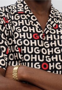 Maglietta nera a maniche corte con un audace motivo testuale bianco e rosso "HUG", dotata di colletto a incavo e taschino frontale con bottone. Orologio d'oro visibile sul polso.