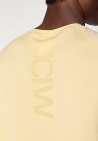 Κίτρινο αθλητικό πουκάμισο με στρογγυλή λαιμόκοψη και υφασμάτινο λογότυπο εκτυπωμένο κατακόρυφα στην πλάτη. Το ύφασμα φαίνεται λείο με ελάχιστες ραφές.