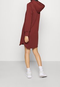 Bordowa sukienka z kapturem ze długimi rękawami, bocznymi kieszeniami i zaokrąglonym dołem. Noszona z białymi sneakersami i skarpetkami. Gładka faktura.