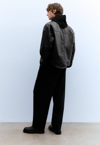 Zwarte leren jas met capuchon, loszittende zwarte broek en zwarte schoenen. De jas heeft een gladde textuur en een minimalistisch ontwerp.