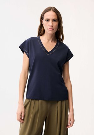 Pitkähiuksinen nainen, jolla on kultaisten korvarenkaiden korut, tummansininen V-kauluksellinen lyhythihainen paita ja oliivinvihreät pliseeratut housut valkoista taustaa vasten.