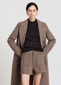 Cappotto marrone a spina di pesce sopra un maglione a trecce marrone scuro, abbinato a pantaloncini marroni strutturati a vita alta con tasche applicate frontali.