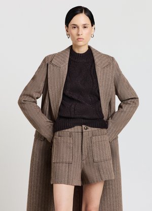 Brauner Fischgrätenmantel über einem dunkelbraunen Pullover mit Zopfmuster, kombiniert mit braunen, strukturierten Shorts mit hoher Taille und aufgesetzten Vordertaschen.