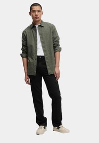 Camisa verde de botones, camiseta blanca, jeans negros y zapatillas beige con detalles verdes. El modelo se encuentra de pie con las manos relajadas a los lados.