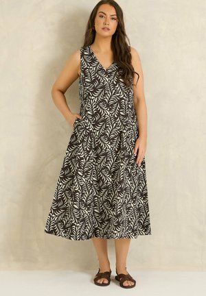 Kobieta w czarno-białej wzorzystej sukience midi bez rękawów z dekoltem w serek i brązowych sandałach, stoi z jedną ręką w kieszeni.