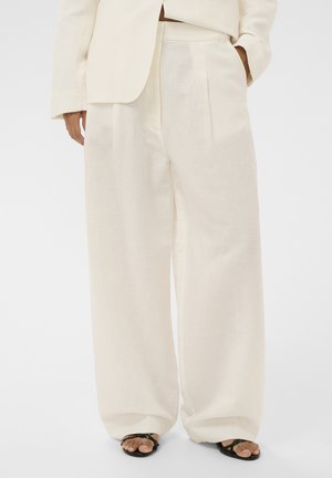 Személy laza, krémszínű nadrágot és hozzá illő blézert visel, fekete nyitott orrú szandállal, kezét a zsebében tartva áll.