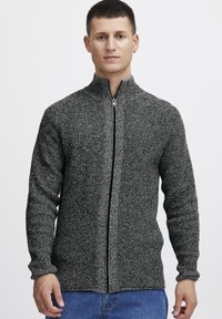 Sudadera gris con cremallera y un diseño de punto texturizado. Presenta un cuello alto y mangas largas, combinada con jeans azules.