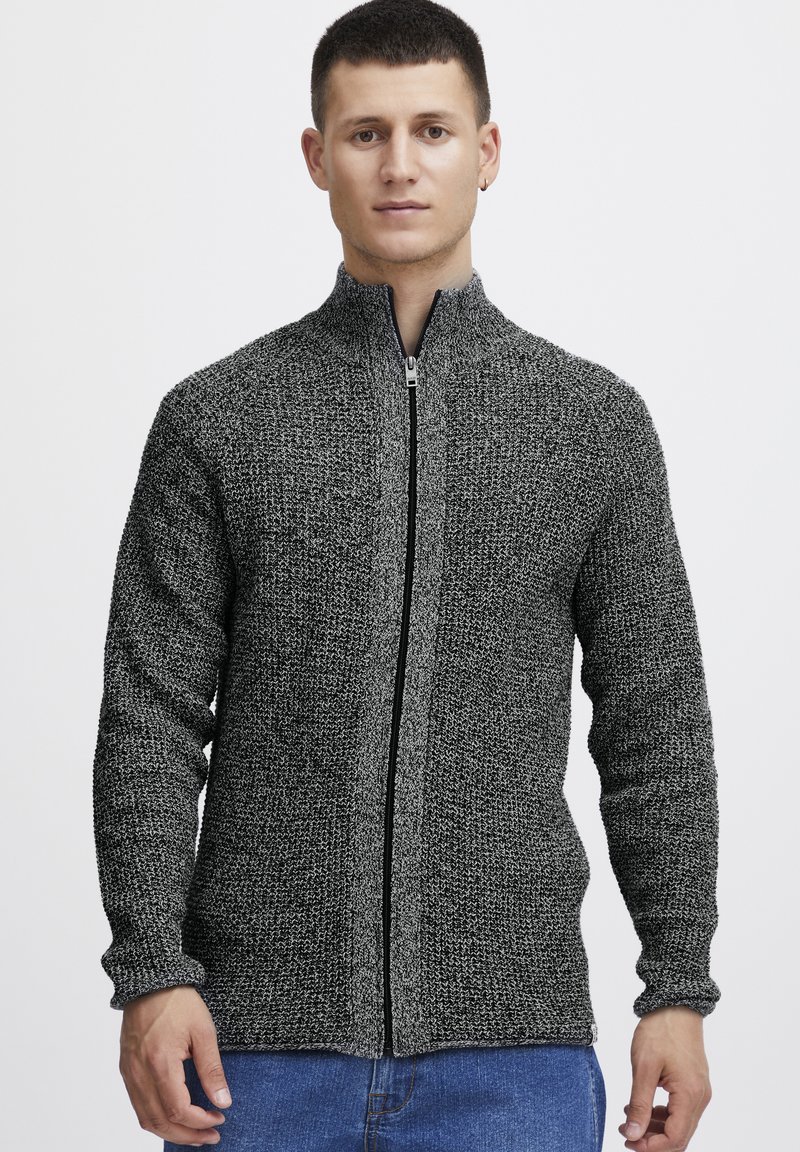 Sudadera gris con cremallera y un diseño de punto texturizado. Presenta un cuello alto y mangas largas, combinada con jeans azules.
