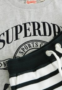 Grå sweatshirt med texten "SUPERDRY" i svart. Nedanför, mörkgröna joggingsbyxor med vita horisontella ränder och snörningsdetaljer.