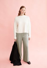 Cremefarbener gestrickter Pullover, weite hellgrüne Hose, schwarze Jacke in der Hand und schwarze Schuhe an einem Model vor einem pinken Hintergrund.