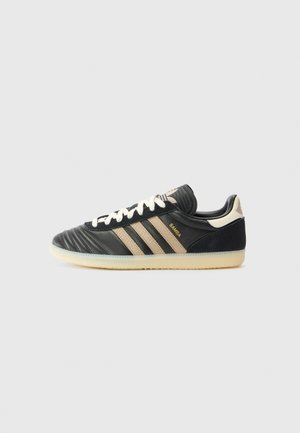 Czarny but sportowy Adidas Samba z beżowymi paskami, białymi sznurowadłami i przezroczystą podeszwą na jasnoszarym tle, widok z boku.