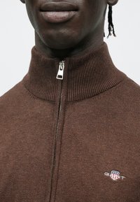 Brūna džemperis ar rāvējslēdzēju, ribota apkakle un teksturēts audums. Aprīkots ar sudraba krāsas rāvējslēdzēju un nelielu GANT logotipu uz krūtīm.