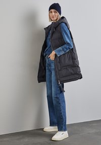 Gilet imbottito nero con cappuccio, indossato sopra una camicia di denim blu e jeans blu, abbinato a sneakers bianche su un pavimento scuro.