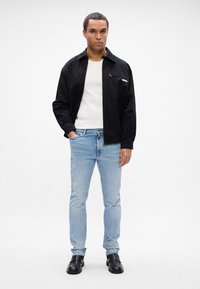 Černá bunda s kapsou na zip, nošená přes bílou košili. Světlé modré džíny a černé boty na slip-on dokončují outfit. Jednoduchý design, pohodlný střih.