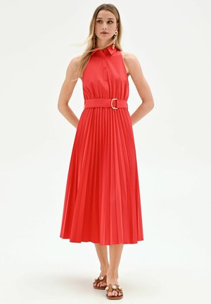 Femme en robe midi plissée rouge sans manches avec une large ceinture et une boucle dorée, portant des boucles d'oreilles remarquables et des sandales ornées de bijoux.